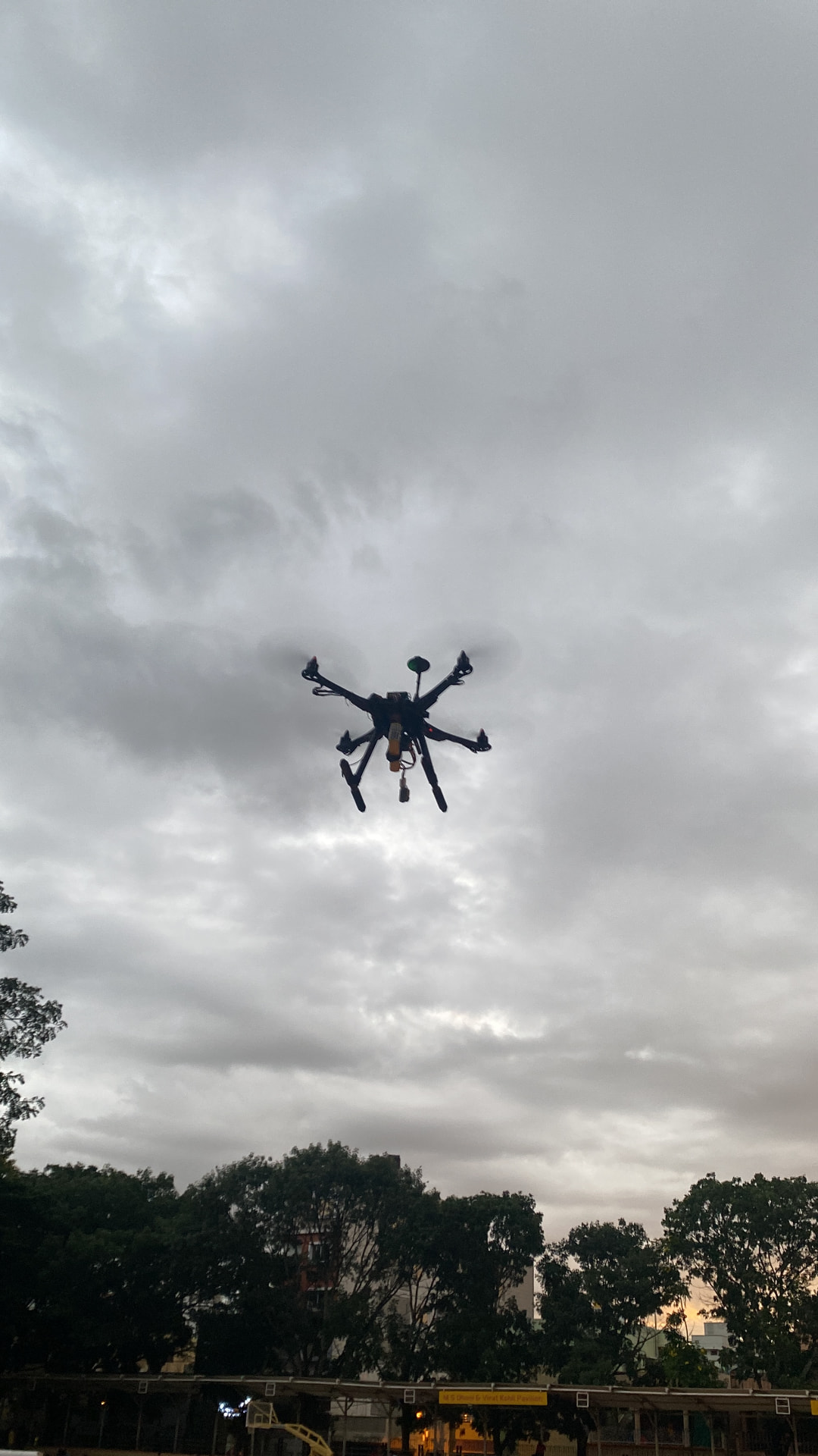 A quadcopter drone in flight against an overcast sky, seen from below, representing autonomous low-light aerial surveillance.